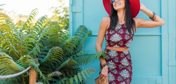 stylish beautiful woman in red hat posing on blue background, printed outfit, summer style, fashion trend, top, skirt, skinny, straw handbag, sunglasses, accessories, smiling, happy, tropical vacation